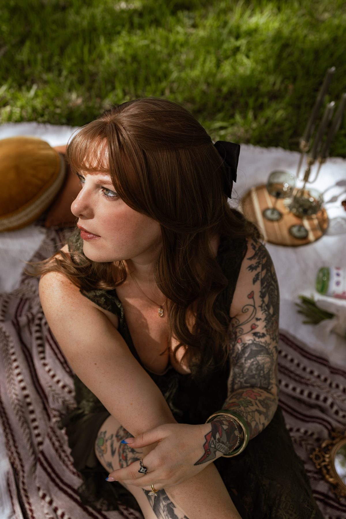 A woman in a dark lace dress sits on a patterned blanket at a styled outdoor picnic, looking away into soft daylight.